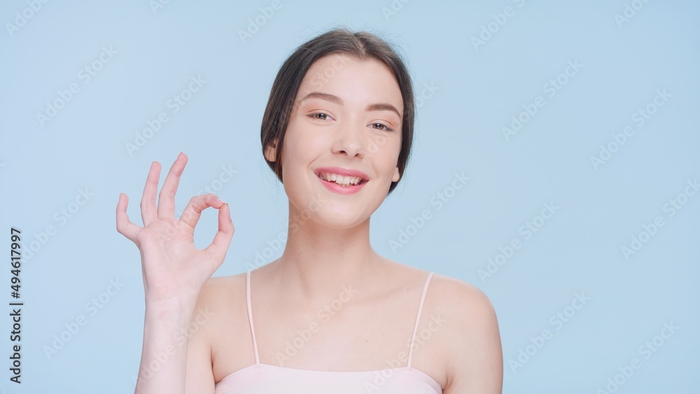 Close-up portrait of young attractive brown-haired woman with a perfect skin make an ok hand gesture, looks at the camera and smiles on blue background | ok sign Close-up portrait of young attractive brown-haired woman with a perfect skin make an ok hand gesture, looks at the camera and smiles on blue background | ok sign