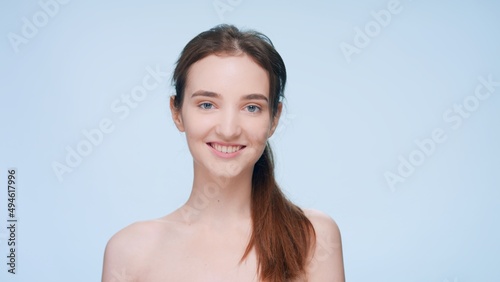 Close-up beauty portrait of young brunette woman with a ponytail looks at camera and smiles against blue background | Skin care concept photo