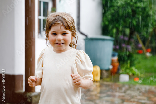 Little preschool girl running through heavy summer rain in garden. Happy smiling wet toddler child having fun with splashing and jumpin in puddles. Activity for children on rainy weather day. photo