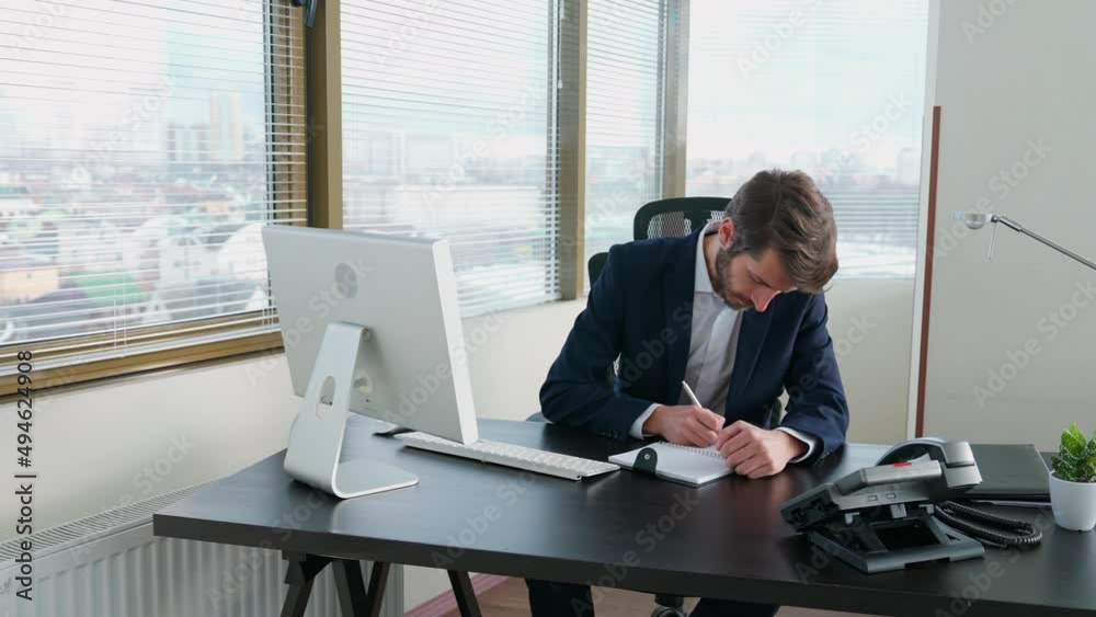 Young businessman working at a computer sitting at work desk, workaholic at work, manager writes the information in a notebook, office with panoramic windows. Young businessman working at a computer sitting at work desk, workaholic at work, manager writes the information in a notebook, office with panoramic windows.