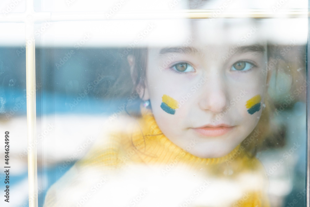 A sad child look at the window with the flag of Ukraine painted on the cheek, worries and fear. Humanitarian aid to children, world peace, security.