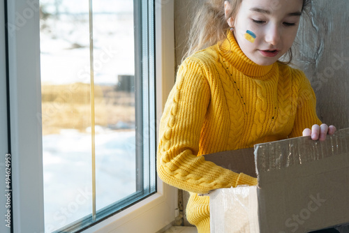 A child on the window with a humanitarian aid box with the flag of Ukraine painted on his cheek. A box with personal belongings, moving, refugee, emigrant