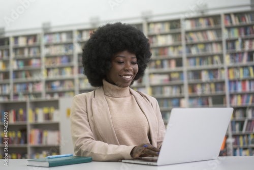 Young smiling African student using laptop computer studying sitting in modern library, education concept. Beautiful Nigerian woman copywriter typing on keyboard working online at workplace 