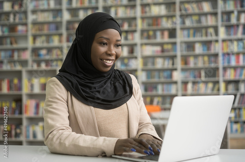 Young smiling African student wearing hijab using laptop computer studying sitting in modern library, education concept. Beautiful happy Nigerian woman typing on keyboard working online at workplace photo