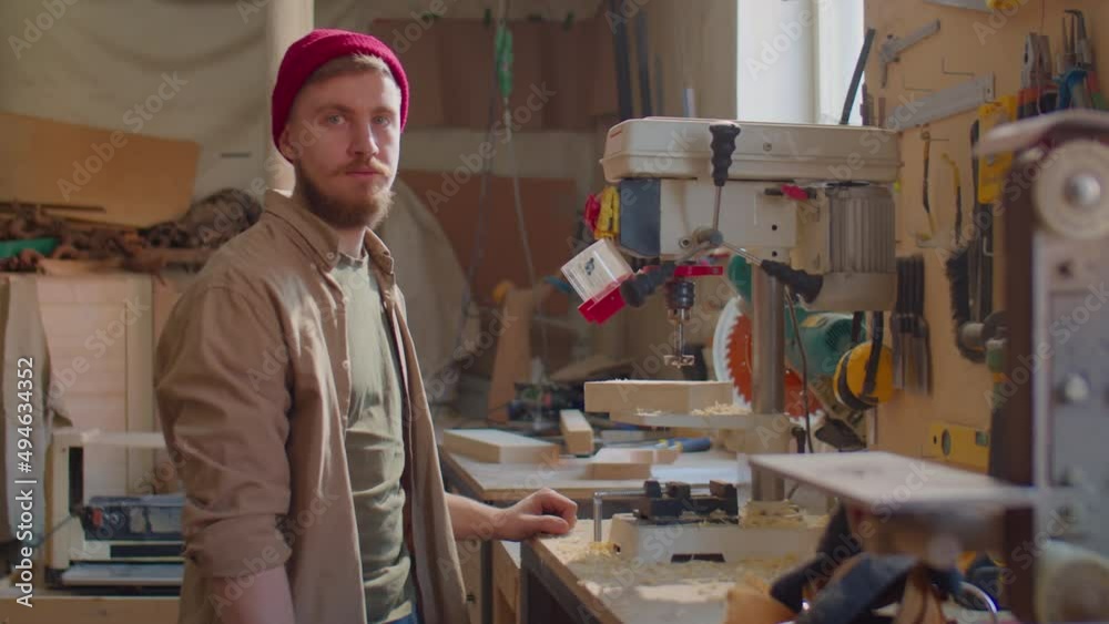 Young bearded carpenter in red beanie hat blowing sawdust from wooden plank and posing for camera in woodworking workshop Young bearded carpenter in red beanie hat blowing sawdust from wooden plank and posing for camera in woodworking workshop