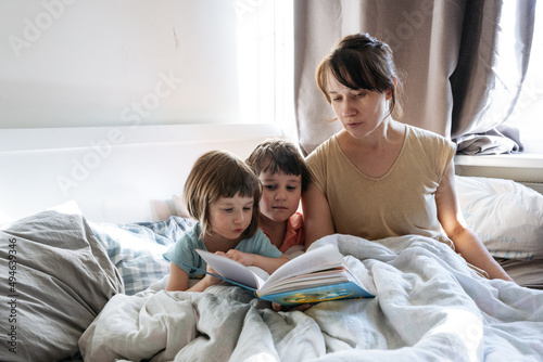 Woman reading a book to siblings while they are all sitting in the bed © Maria
