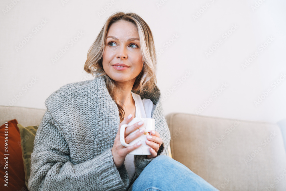 Young beautiful woman forty year with blonde long curly hair in cozy knitted grey sweater with cup of tea in hands in bright interior at home Young beautiful woman forty year with blonde long curly hair in cozy knitted grey sweater with cup of tea in hands in bright interior at home