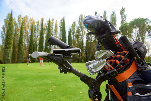 Fototapeta Naklejka Na Ścianę i Meble -  Golf cart with bag and golf clubs in the fairway of a golf course, parked at the edge of the green. In the background the players are preparing to pocket the ball