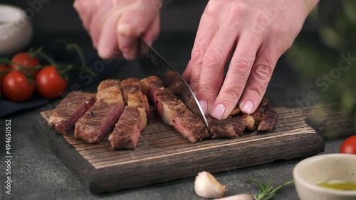 Chef with Knife Cuts of Grilled Beef Steak on Rustic Cutting Board on Gray Background. Concept of Delicious Meat Food. Juicy Sliced Rib Eye Steak with Salt. 