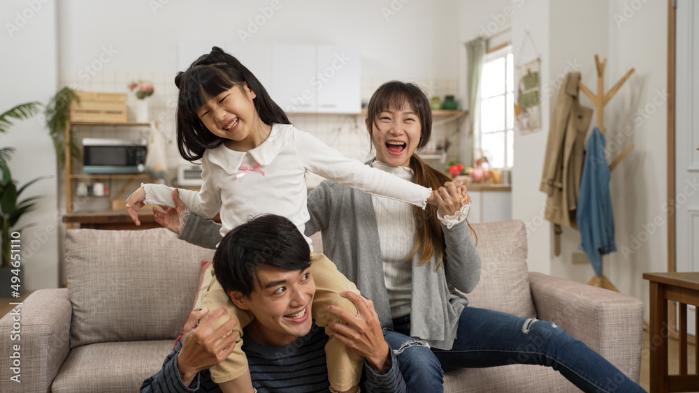 loving asian father giving his daughter piggyback ride on shoulders with mother’s support. the girl laughs with excitement while playing with family at home loving asian father giving his daughter piggyback ride on shoulders with mother’s support. the girl laughs with excitement while playing with family at home