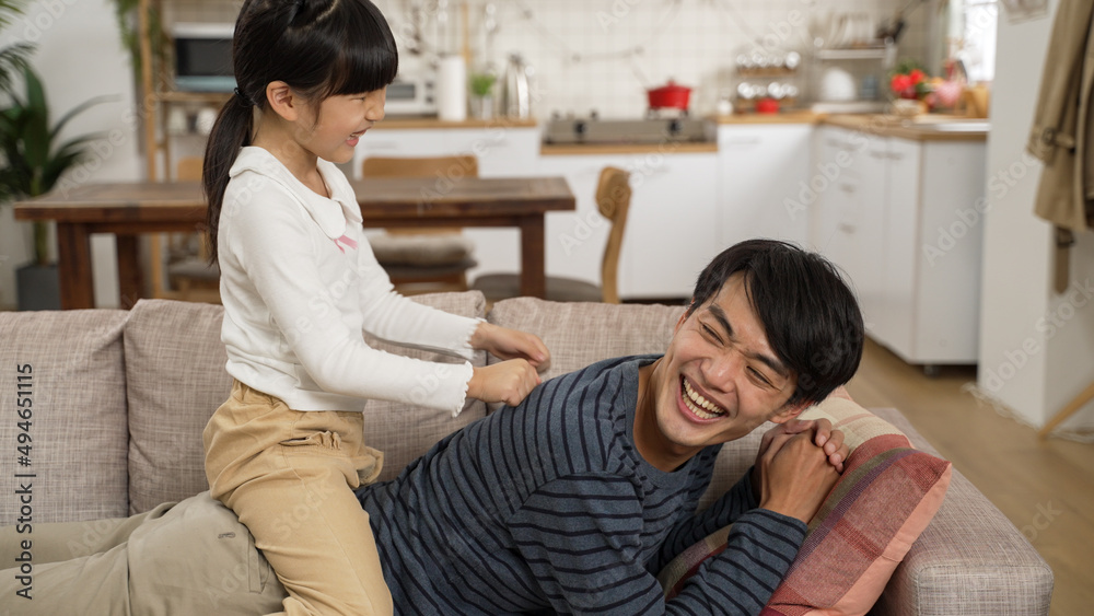 cheerful asian little girl sitting on her dad’s back giving him a massage at home. the father lying prone on the sofa and telling his daughter to punch harder.