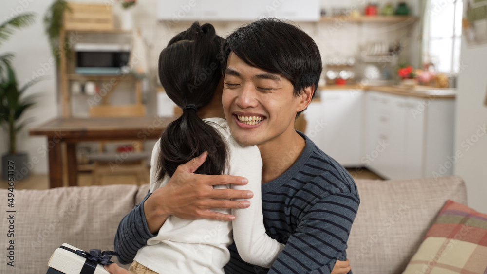 view from behind Asian little girl walking up to her dad and showing him a surprise gift in living room at home. happy father embracing her daughter for the sweet act on father's day view from behind Asian little girl walking up to her dad and showing him a surprise gift in living room at home. happy father embracing her daughter for the sweet act on father's day