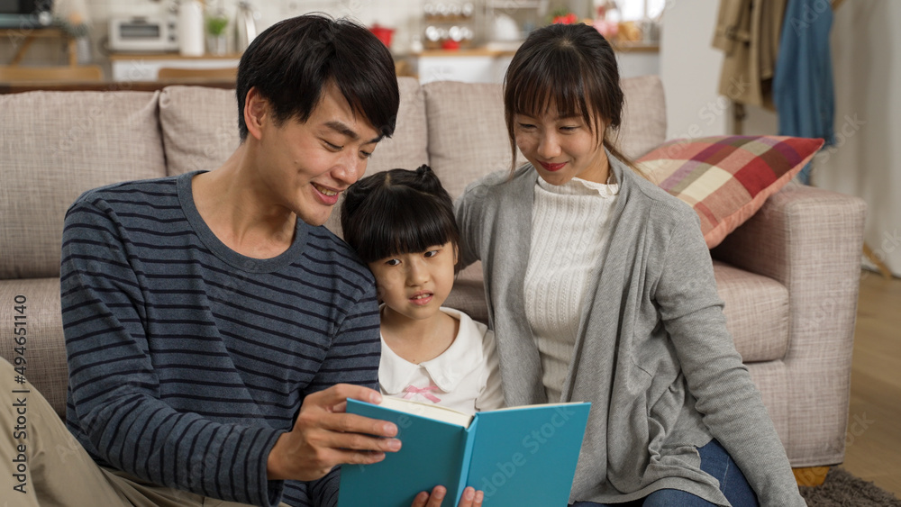 cheerful asian family of three enjoying reading together in the living room at home. the smiling father is having fun telling story with a book in hand.