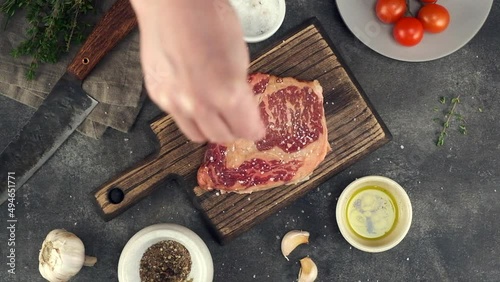 Chef sprinkling salt and pepper on ribeye steak, top view
