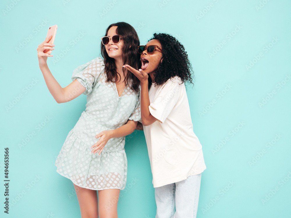 Two young beautiful smiling international hipster female in trendy summer clothes. Sexy carefree women posing near blue wall in studio. Positive models having fun in sunglasses. Taking selfie photos Two young beautiful smiling international hipster female in trendy summer clothes. Sexy carefree women posing near blue wall in studio. Positive models having fun in sunglasses. Taking selfie photos
