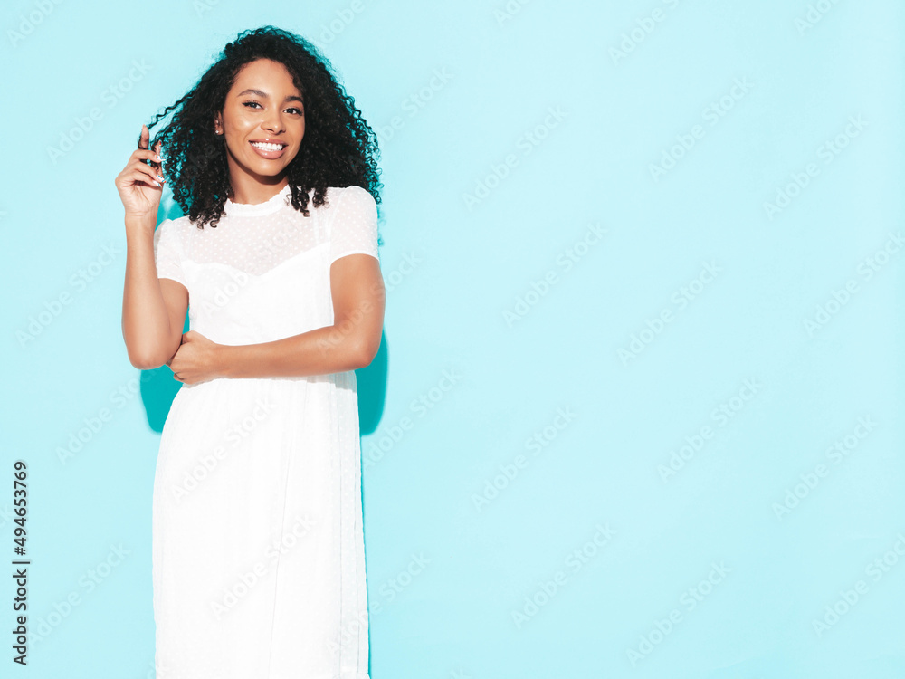 Beautiful black woman with afro curls hairstyle. Smiling model dressed in white summer dress. Sexy carefree female posing near blue wall in studio. Tanned and cheerful. Isolated Beautiful black woman with afro curls hairstyle. Smiling model dressed in white summer dress. Sexy carefree female posing near blue wall in studio. Tanned and cheerful. Isolated
