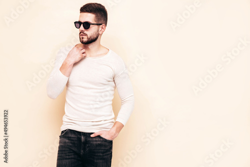 Portrait of handsome confident stylish hipster lambersexual model.Man dressed in white sweater and jeans. Fashion male posing near wall in studio in sunglasses
