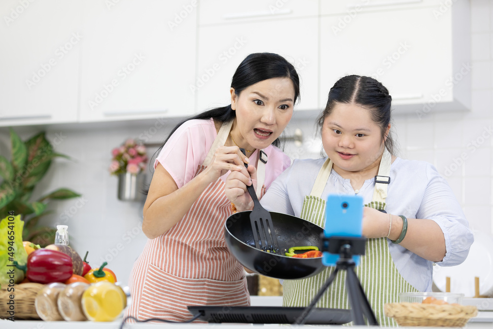 mother and down syndrome teenage girl or her daughter cooking food together and live streaming online via smartphone on tripod in a kitchen