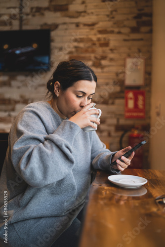 Obraz na płótnie Chica joven guapa tomando té en una cafetería muy acogedora con sudadera gris y mirando el smartphone w salonie