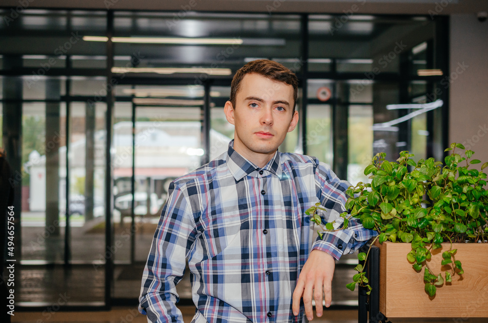 a guy in a shirt leans on a flowerpot and looks at the camera against the background of glass doors a guy in a shirt leans on a flowerpot and looks at the camera against the background of glass doors