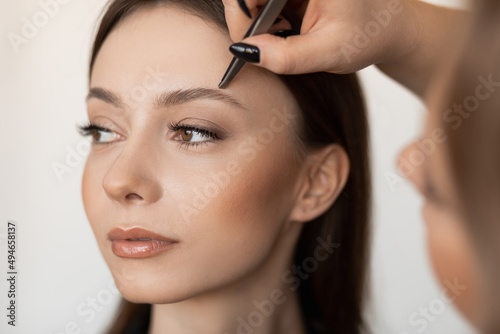 Close-up of young gorgeous woman with long dark hair tweezing eyebrows by professional make-up artist in black clothes. photo