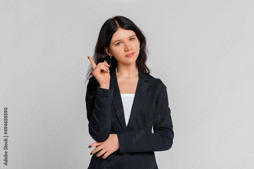 Serious brunette teen girl in black blazer pointing finger up, scolding or telling off someone, says: Attention please, standing over grey background Serious brunette teen girl in black blazer pointing finger up, scolding or telling off someone, says: Attention please, standing over grey background