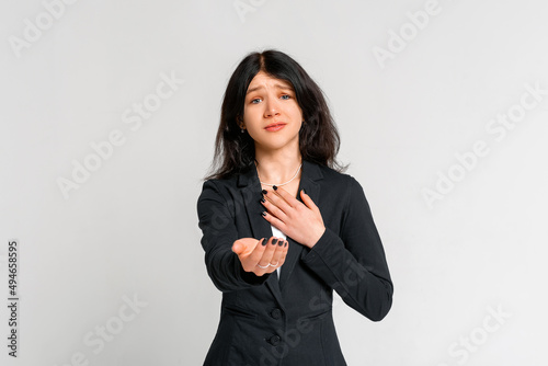 Cute teen girl ask for help with compassionate gaze, reaching hands of hand to the camera, standing in black blazer over grey background. Human emotions concept