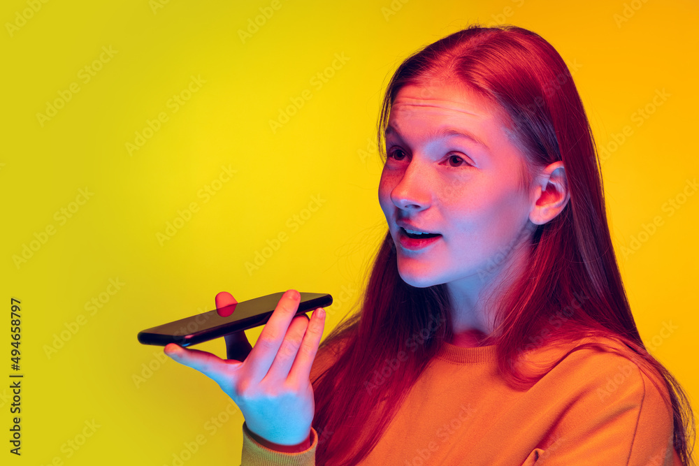 Portrait of teen girl recording voice message on her phone isolated over yellow studio background in neon light