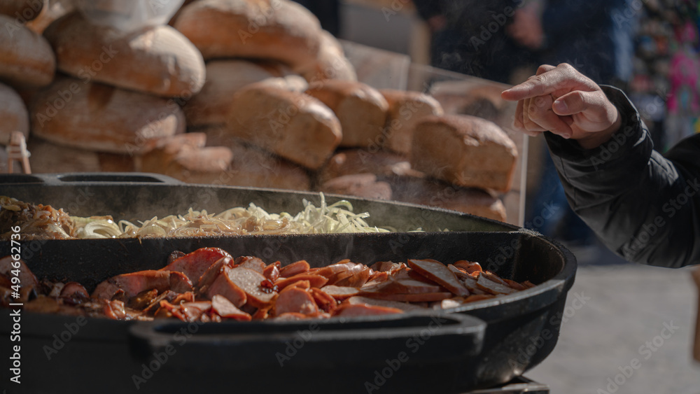 Behind puffs of white steam over sausages and onion rings, which are being fried on an electric grill on a street counter, the hand of the buyer is visible. Street Food Festival at the Easter Market