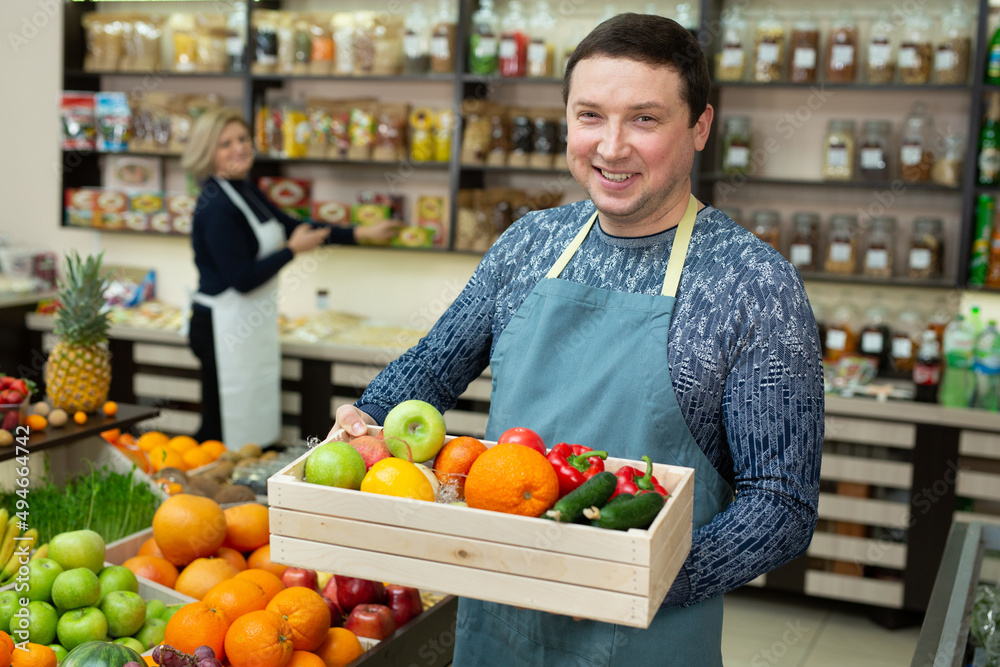 Smiling male salesman holds a wooden box with vegetables and fruits in the store. Smiling male salesman holds a wooden box with vegetables and fruits in the store.
