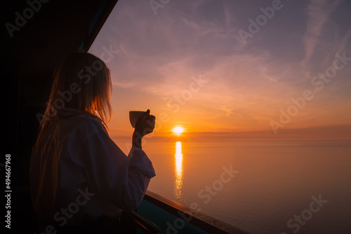 Happy young woman in a white shirt is drinking coffee from a white cup on the balcony, watching the sun go down over the horizon of the sea.Woman with a cup on the background of a beautiful sunset