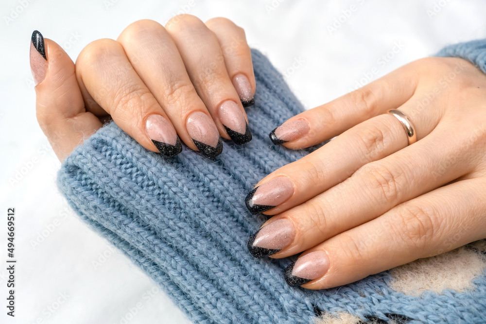 Beautiful French manicure with black contour and sequins. Well-groomed female hands with painted nails with gel polish on a cozy background of blue clothes. Fashionable color of gel nail polish.