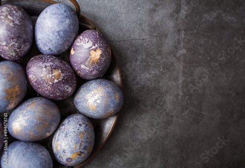 Purple, blue and golden eggs in an iron plate on a dark background. The purple hue trend of 2022 is veryperi. Natural dye karkade tea. Top view.Easter card with a copy of the place for the text. 