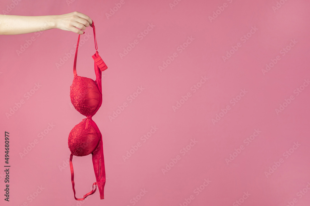 A woman's hand holding a red bra on a pink background. The concept of women's breasts, underwear. Copy space - obrazy, fototapety, plakaty 