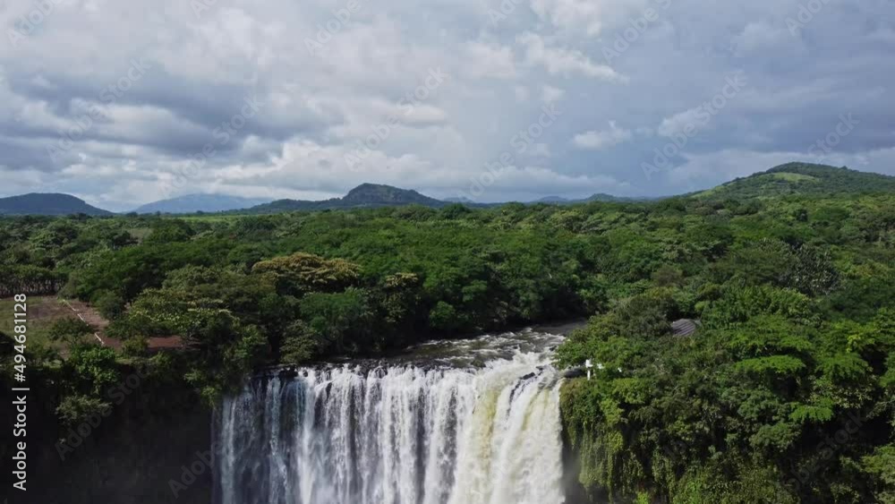 Tilt down shot featuring a beautiful waterfall in the Mexican jungle is surrounded by lush green vegetation on both sides with the view of hills in the background. Tilt down shot featuring a beautiful waterfall in the Mexican jungle is surrounded by lush green vegetation on both sides with the view of hills in the background.
