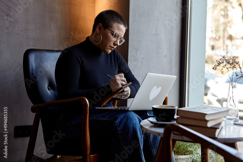 A young girl with glasses, with short hair is sitting in a cafe and working on a laptop. The girl looks at her laptop and makes notes in a notebook. The concept of distance learning