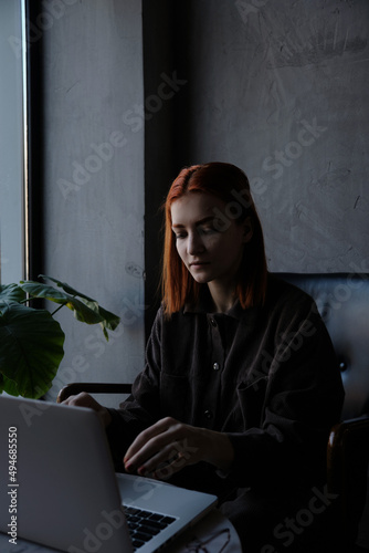 a red-haired freelance woman with glasses works remotely on a laptop in a cafe.x