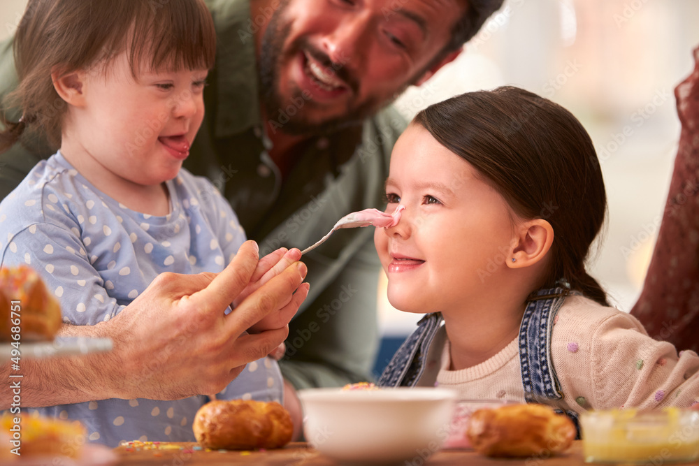 Family With Down Syndrome Daughter Baking And Decorating Cakes Sitting Around Table At Home Family With Down Syndrome Daughter Baking And Decorating Cakes Sitting Around Table At Home