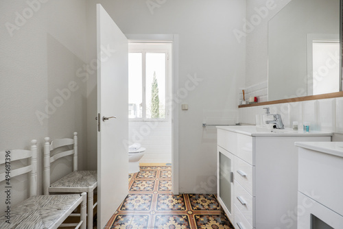 Bathroom with twin white porcelain sinks, frameless mirror on the wall, and cement tile floors with matching white chairs