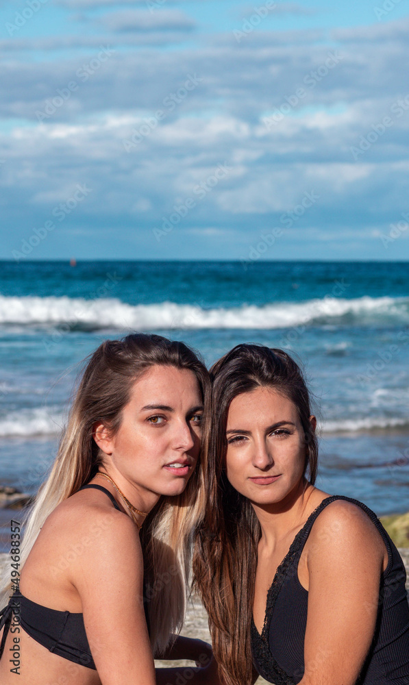 Two young blonde and brunette women looking at the camera at the beach on bikini