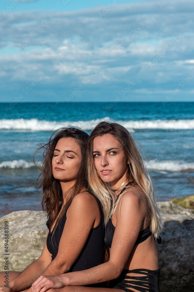 Two lesbian women hugging on the beach in bikinis between rocks and ocean