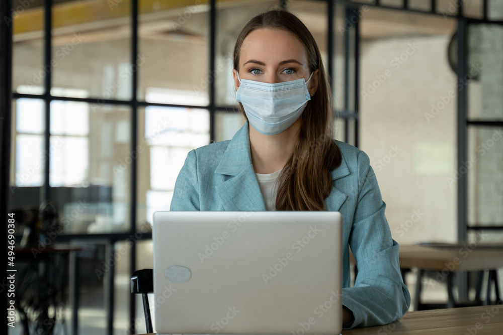 Young woman puts a medical mask on her face and works at a laptop in the office. 