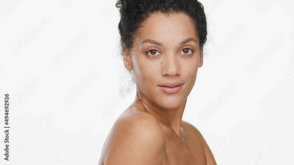 Close-up beauty portrait of attractive young slim African American woman with dark curly hair with bare shoulders looks at the camera over her shoulder on white background | Body care concept