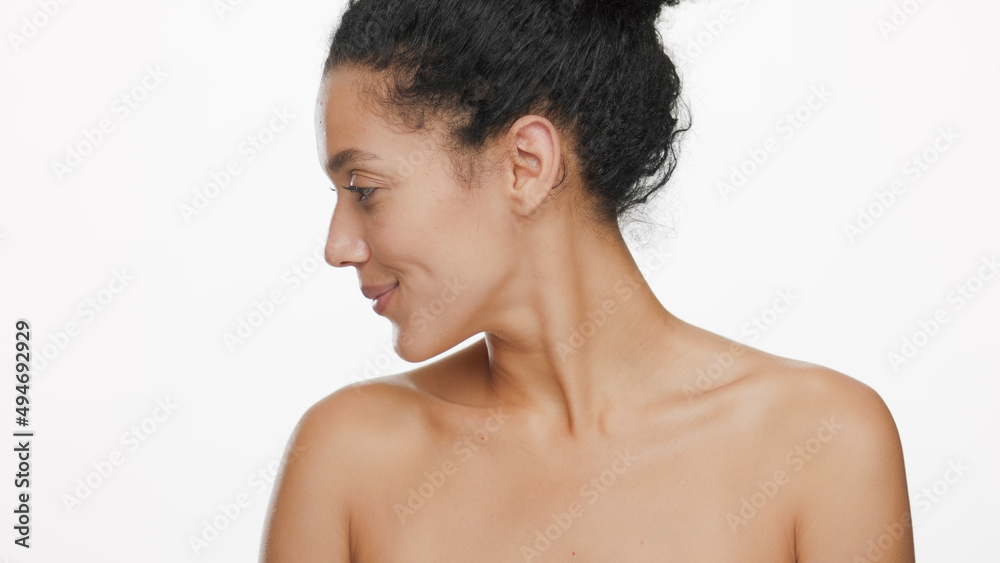 Close-up beauty portrait of young slim pretty African American woman with black curly hair with nude shoulders turns her head to her right shoulder on white background | Skincare concept