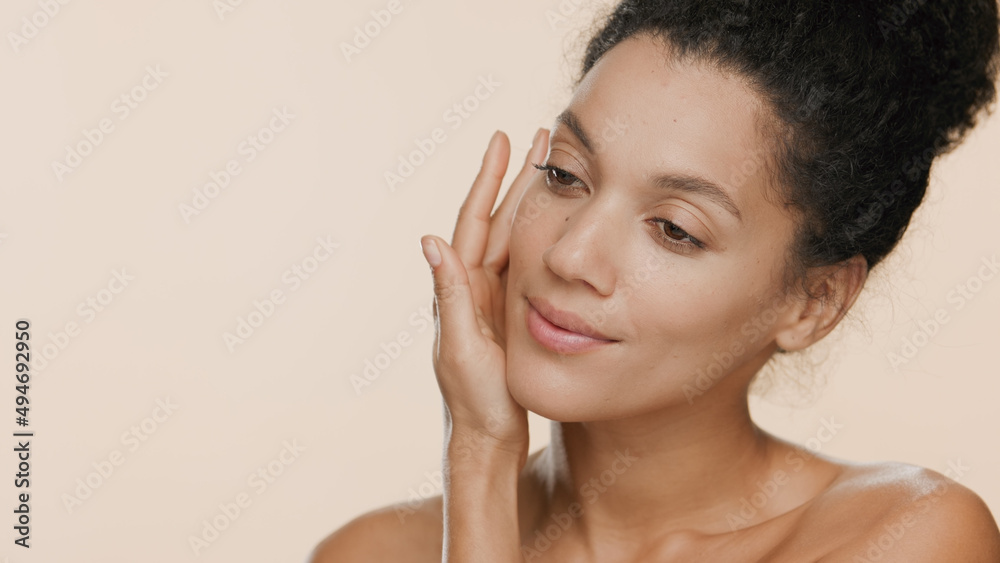 Young slim pretty black-haired African American woman with naked shoulders strokes her face with palm of her hand against beige seamless background | Smooth face concept