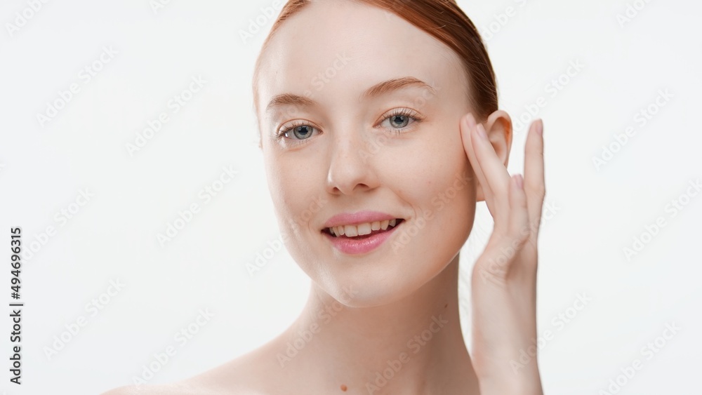 Young positive ginger woman touches her cheek with her finger against light-grey background | Skin care commercial concept