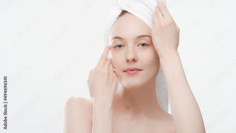 Beautiful young slim European woman with a towel on her head touches her face gently with both hands on white background | Face care commercial concept