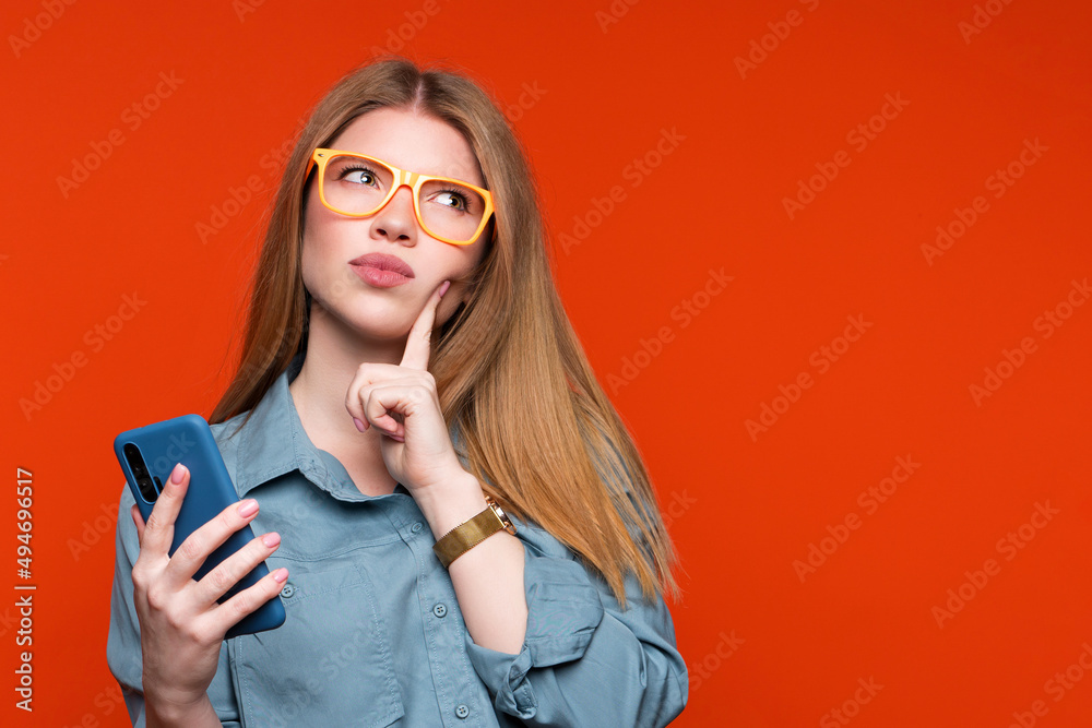 Thoughtful and prudent girl in basic blue shirt and orange glasses with cellphone. Young woman thought about something and looks away with finger to cheek. Online shopping and reasonable consumption.