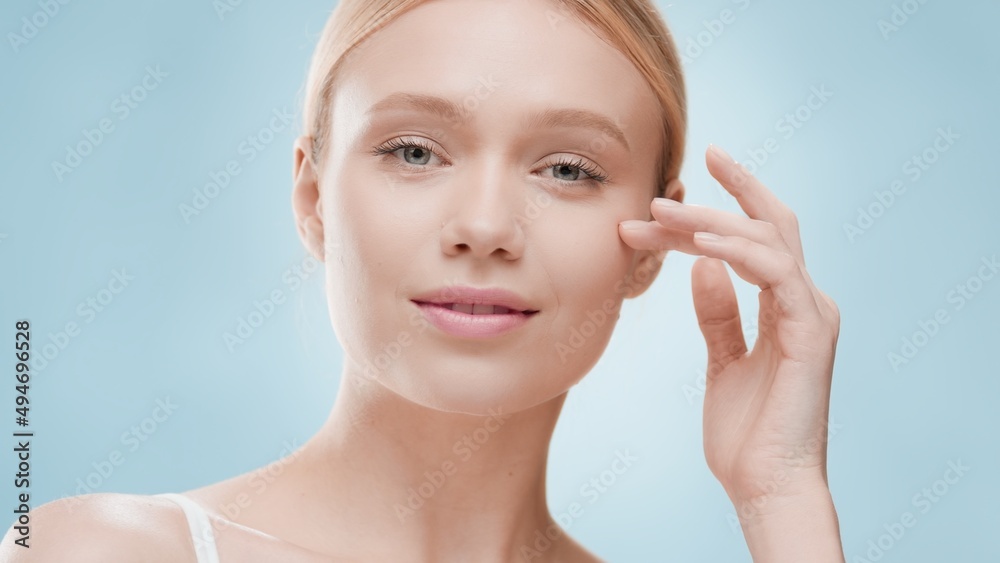 Fototapeta premium Young fit attractive European woman with light hair touches her cheek looking at the camera against blue background | Acne prevention concept