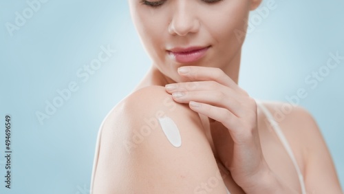 Young slim cute European blond woman applies moisturizing cream on her shoulder against blue background | Moisturizer putting on shot for skincare commercial